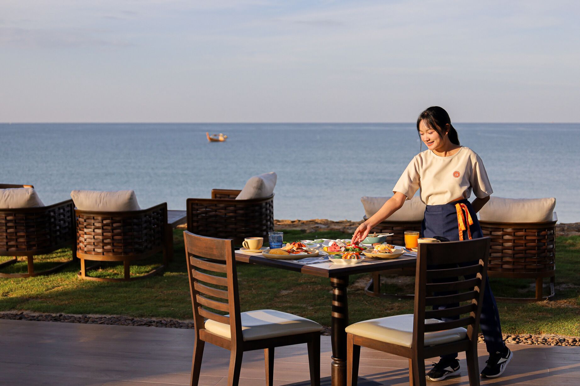 A dining table set outdoors with wicker chairs, a person serving food, and a view of the sea.