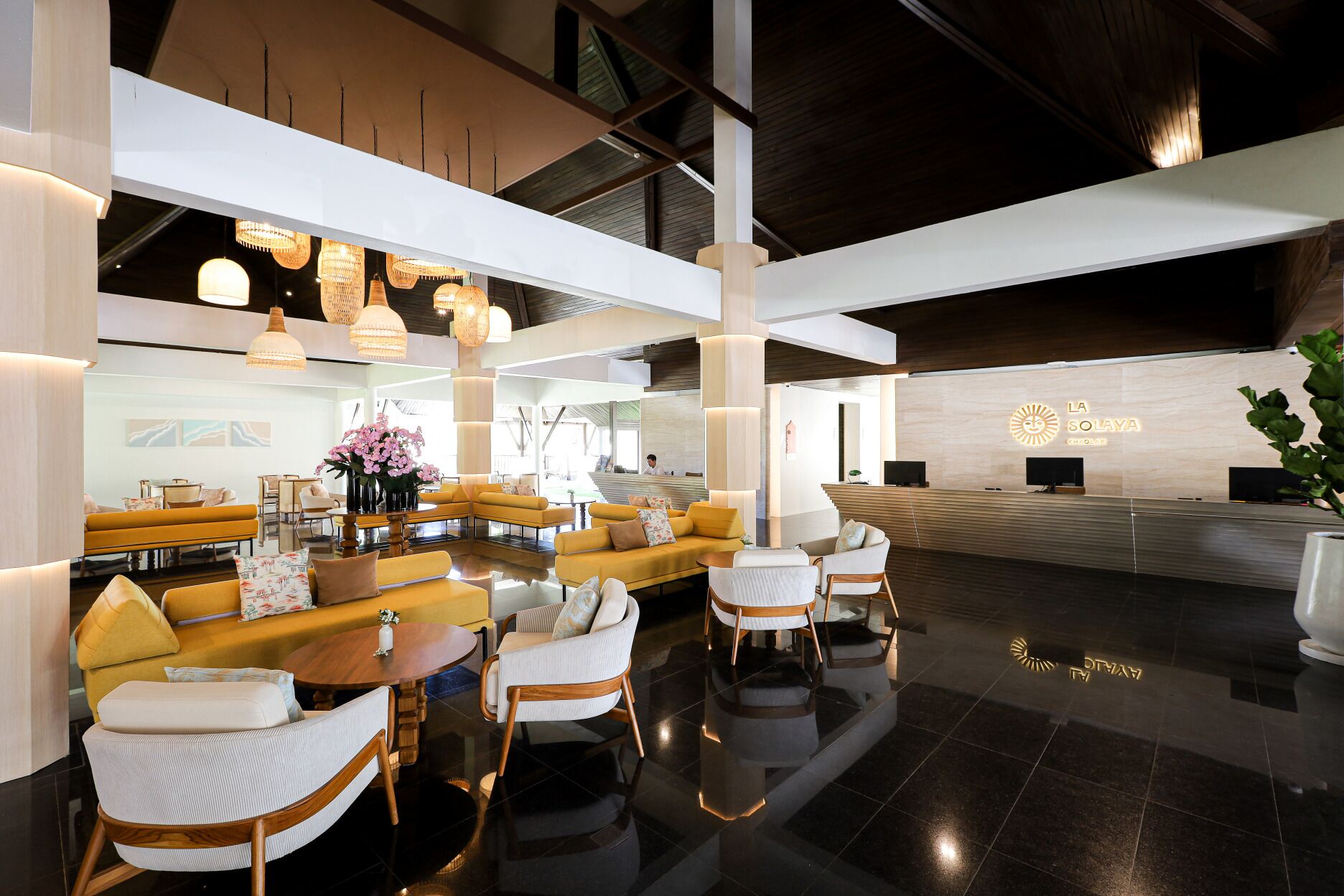 Eine moderne Hotelhalle mit einer Rezeption, einer Sitzecke mit Sofas und Sesseln sowie dekorativer Beleuchtung.