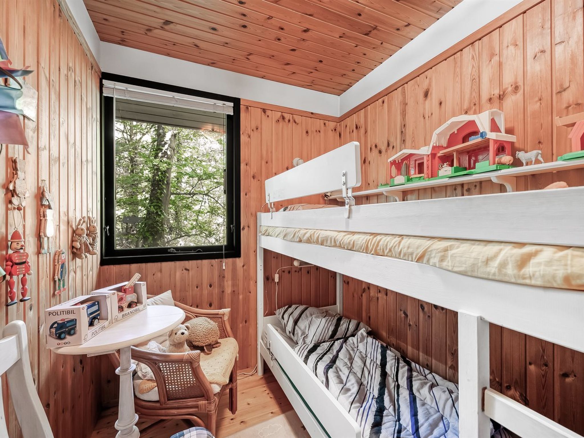 A cozy room with wooden walls, a bunk bed, a small desk with toys, and a window with a view of trees.