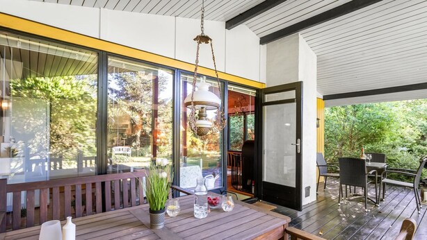 Un área de comedor moderna al aire libre con una mesa y bancos de madera, rodeada de puertas y ventanas de vidrio que dan vistas al jardín.