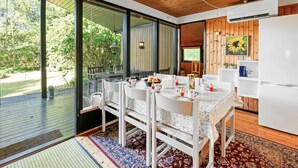 Una cocina moderna con techo de madera y muebles de cocina blancos, un área de comedor con sillas blancas y una mesa dispuesta para cuatro personas, y ventanas amplias que ofrecen vista a la vegetación exterior.