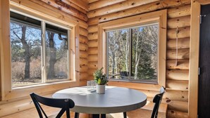 A wooden cabin interior with a round table and two chairs, large windows, and a view of trees outside. - Bear Creek Haven  (Big Bear Lake)