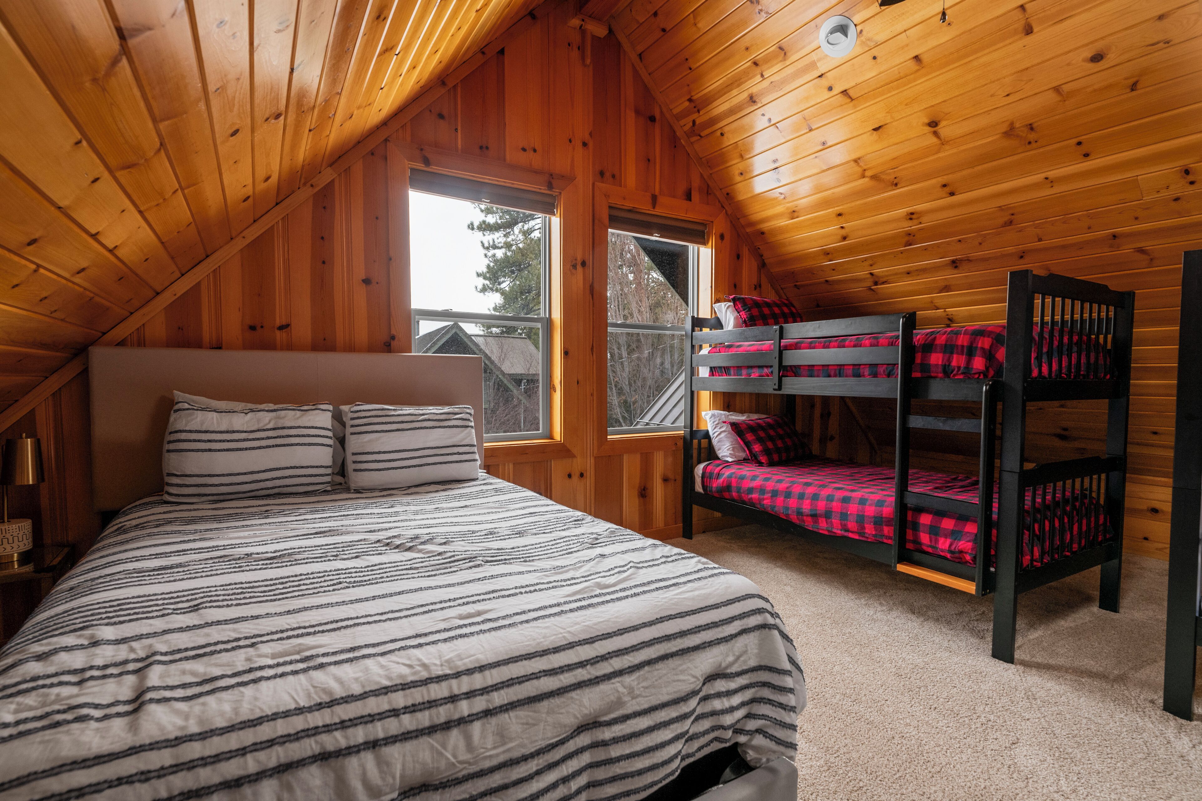 Un dormitorio acogedor en el ático con una cama litera y una cama individual, paredes de madera y un ventanal con vistas al exterior.