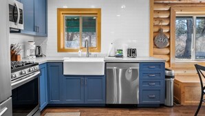 A modern kitchen with blue cabinets, a stainless steel dishwasher, and a wooden window frame. - Bear Creek Haven  (Big Bear Lake)