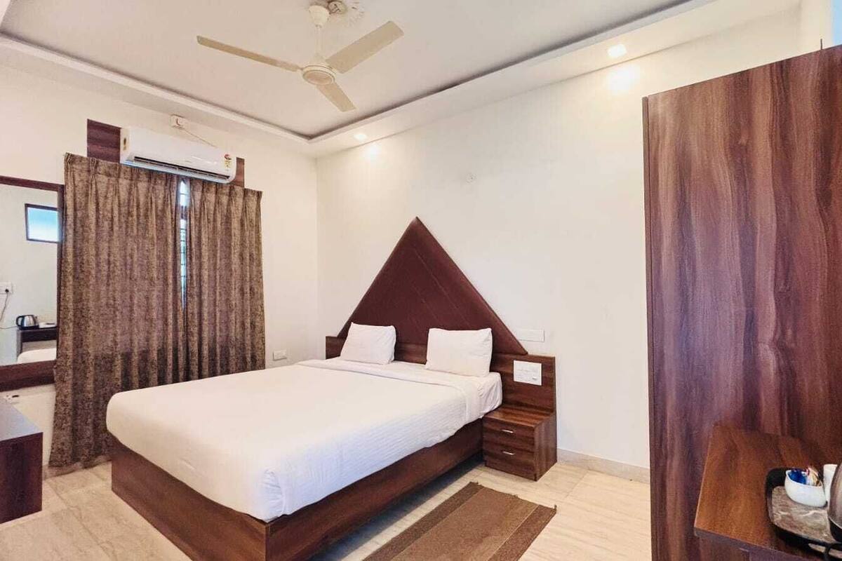 Une chambre d’hôtel comprenant un lit, une tête de lit en bois, une table de chevet, un bureau, une chaise, un miroir, une fenêtre avec des rideaux et un appareil de climatisation.