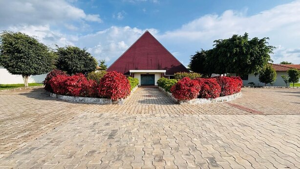 Un edificio con techo triangular y un acceso pavimentado con arbustos florecientes de color rojo.