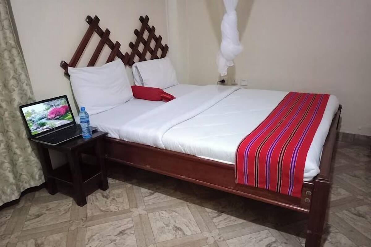 Una cama bien hecha con sábanas blancas y una manta roja a rayas, una laptop sobre una mesita auxiliar y una botella de agua.