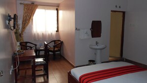 Habitación de hotel con una cama, un escritorio con silla, una mesita con otra silla, un lavamanos y una ventana con cortinas.