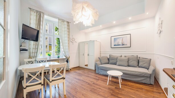 A living room with a wooden floor, white furniture, a grey sofa, a small white table, a TV on the wall, and a window with curtains. - Apartamenty Sun & Snow Przy Monte Cassino (Sopot)