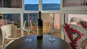 Una mesa con una botella de vino y dos copas, una silla blanca, y vistas al exterior a través de amplios ventanales.