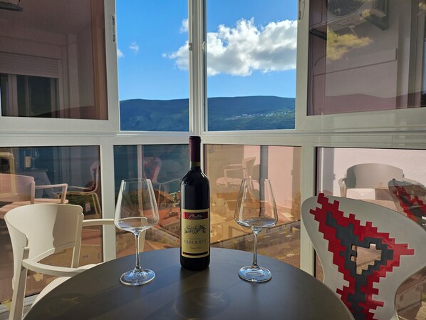 A table with a bottle of wine and two glasses, a white chair, and a view of the outdoors through large windows. - Charming 1-bedroom apartment with WiFi and AC in picturesque Herceg-Novi (Herceg - Novi)