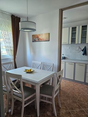 A dining area with a white table and chairs, a painting above the table, and a kitchen in the background.