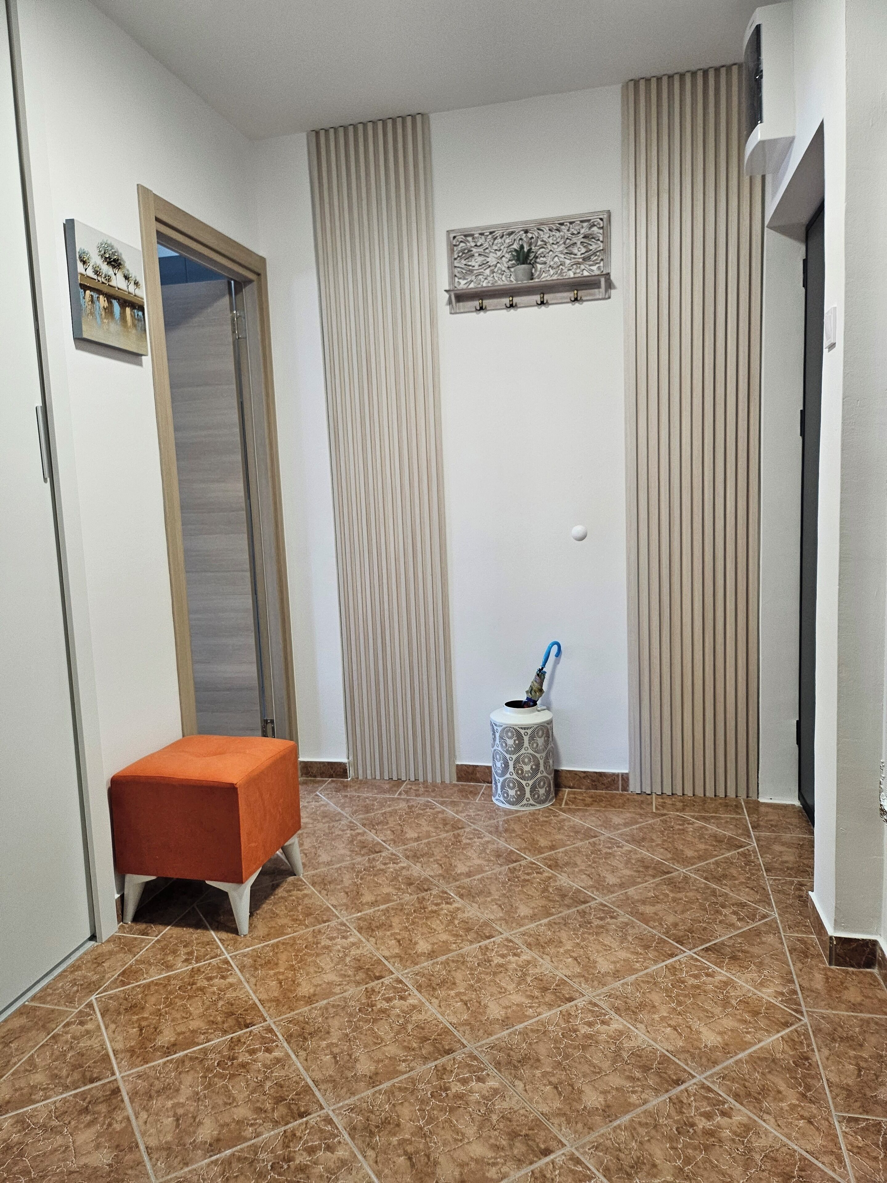 Une pièce avec un sol carrelé, un petit banc orange, une poubelle et une porte dotée d’un miroir.