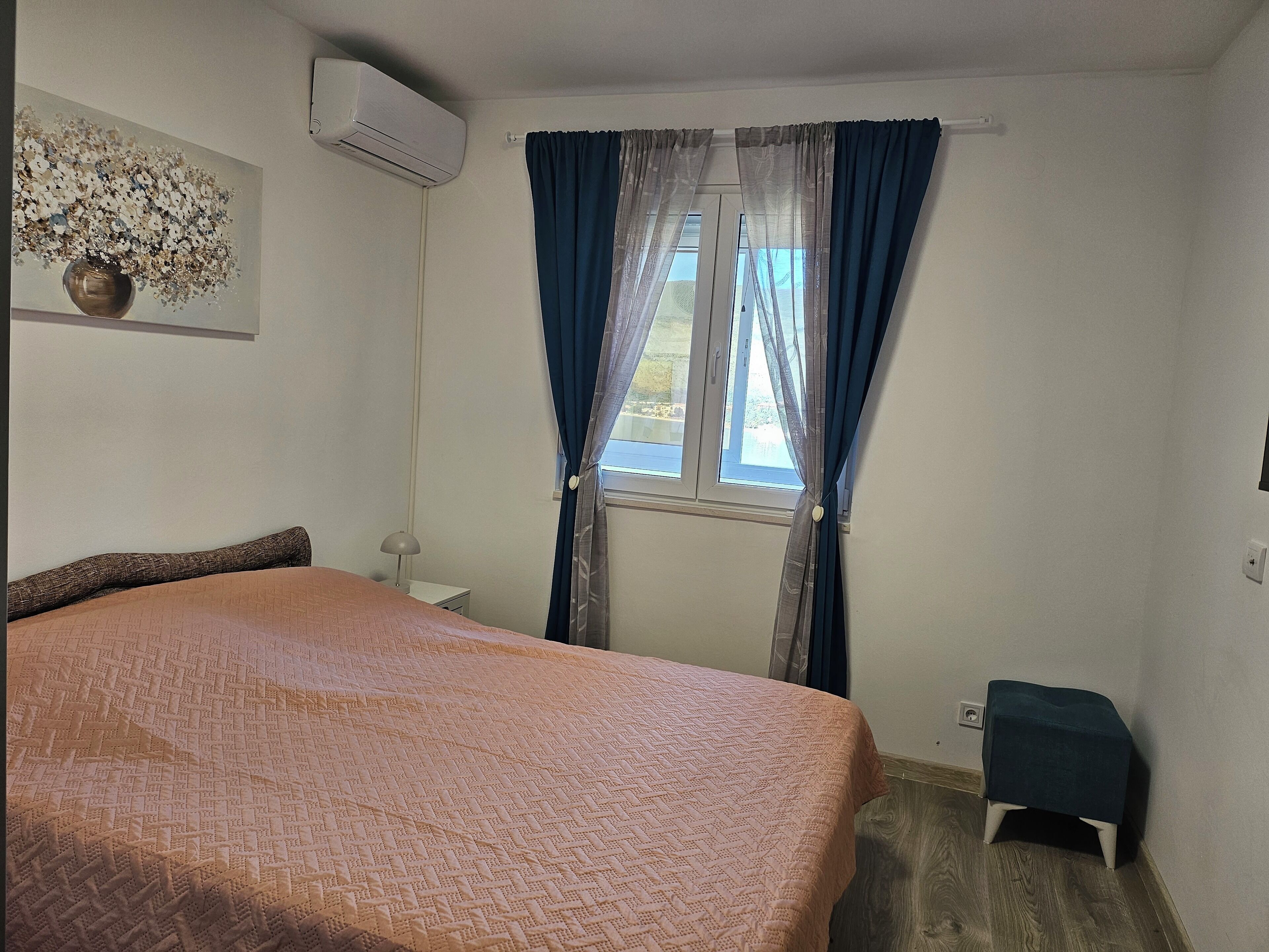 Een slaapkamer met een bed, een nachtkastje, een lamp, een raam met gordijnen en een airconditioner.