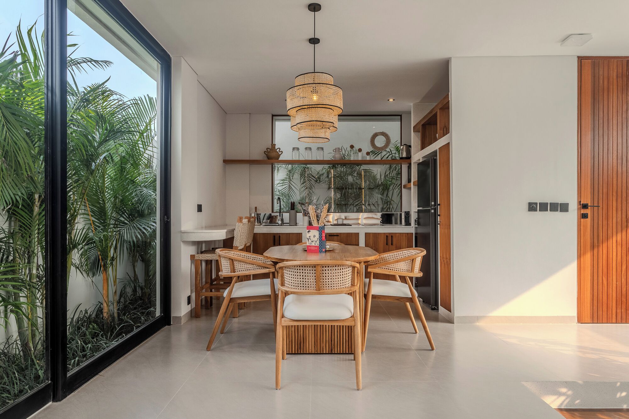 Un espace repas moderne avec une table ronde et quatre chaises en osier, un luminaire suspendu et une vue sur l’extérieur grâce à de grandes fenêtres.