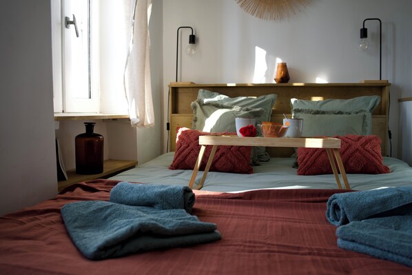 Una habitación con cama, cabecera de madera, mesita de noche, ventana con cortinas y lámpara de pared.