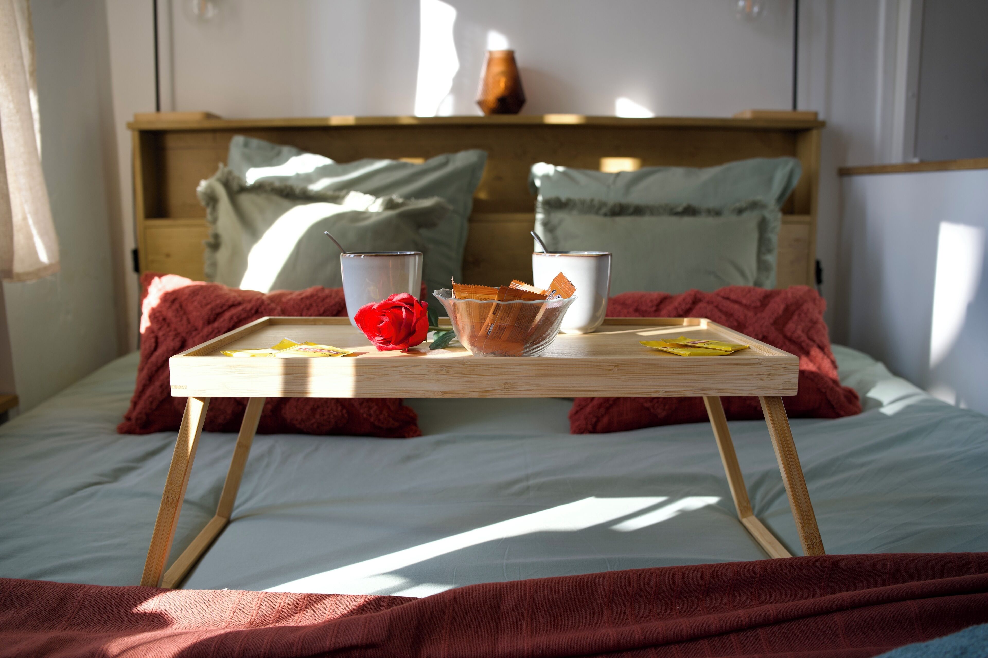 A neatly made bed with a wooden bedside table, a red rose, and a tray with a cup and a bowl of snacks.