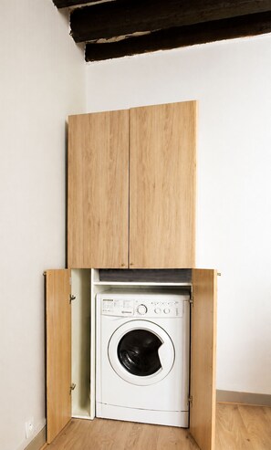 A compact, white washing machine in a small room with wooden cabinetry above and below.
