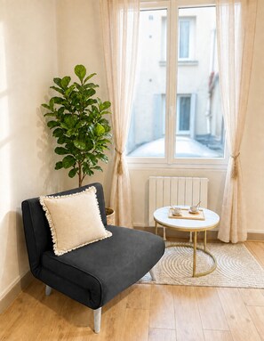 A small, dark-colored sofa with a white cushion, a potted plant, a small round table with a tray and cups, and a window with curtains.