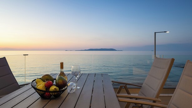 A wooden table with a fruit basket, a bottle, and a glass on a balcony overlooking the sea.