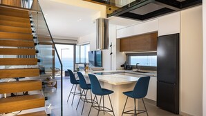 A modern kitchen with a central island, bar stools, a stainless steel refrigerator, and a glass staircase.