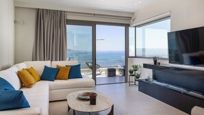 A modern living room with a sofa, coffee table, and a TV. It features large windows with a view of the sea.
