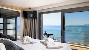 A hotel room with a large window offering a sea view, a bed with neatly arranged towels, and a flat-screen TV mounted on the wall.