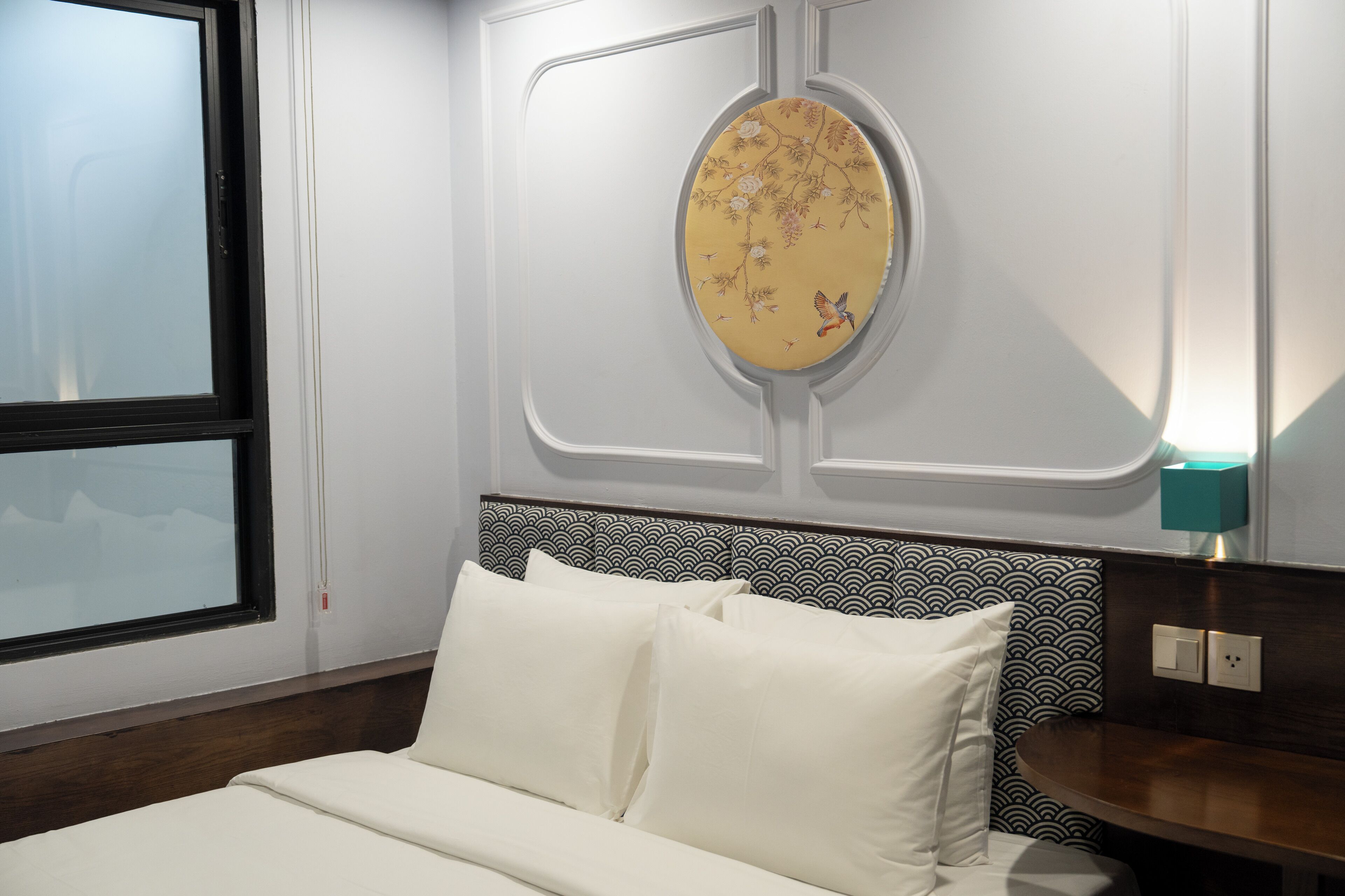 A hotel room with a bed, a decorative wall hanging, a patterned headboard, and a bedside table with a lamp.