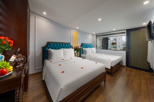 A hotel room with two beds, wooden flooring, a wooden dresser with fruit and a bottle, and a large window with a view of buildings.