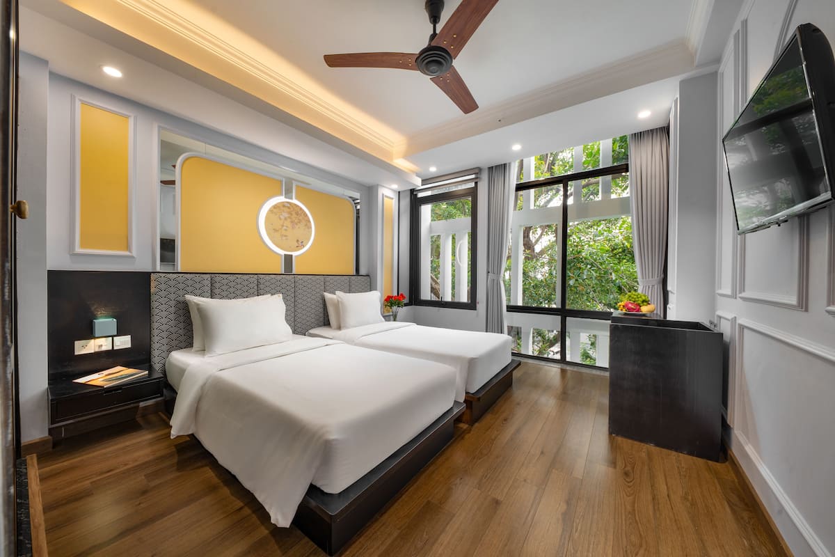 A modern hotel room with two beds, a ceiling fan, a TV, and a large window with a view of greenery.