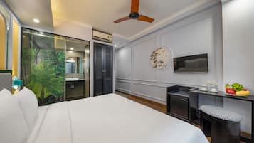 A hotel room with a bed, a desk with a chair, a ceiling fan, a round wall decoration, and a small table with fruits.