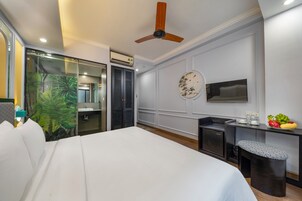 A hotel room with a bed, a desk with a chair, a ceiling fan, a round wall decoration, and a small table with fruits.