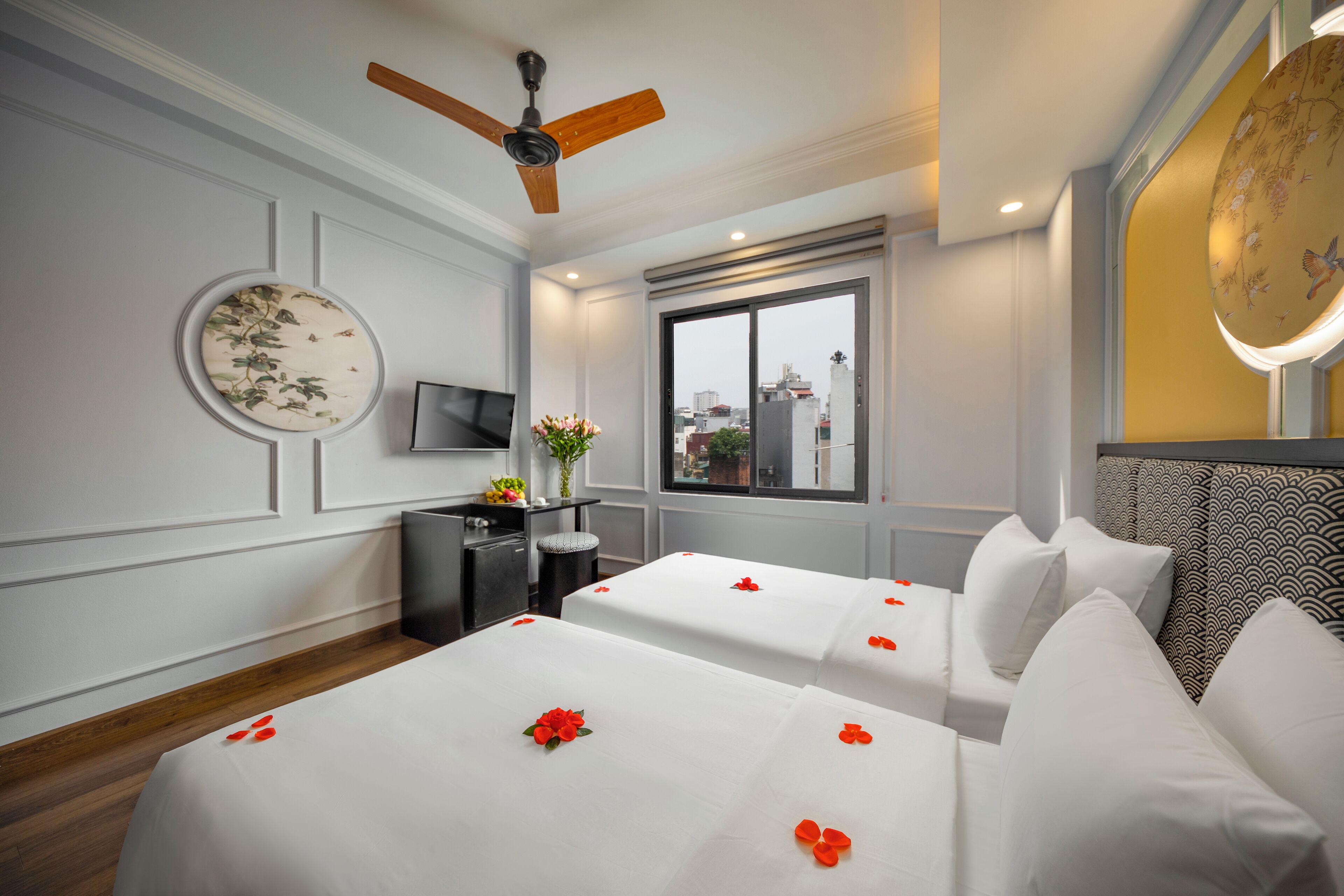 A hotel room with two beds, a ceiling fan, a TV, a window with a city view, and a round mirror.
