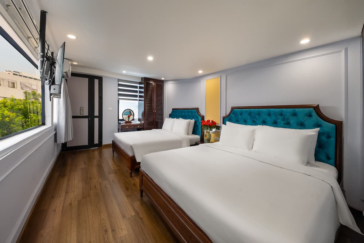 A hotel room with two beds, a wooden floor, a window with blinds, and a view of greenery outside.