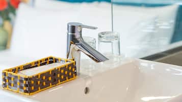 Ein Badezimmer mit einem weißen Waschbecken, einem chromfarbenen Wasserhahn und einem Spiegel, der ein Bett mit blauen Kissen reflektiert.