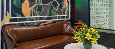 A room with a brown leather sofa, a round table with a vase of yellow flowers, and a large abstract painting on the wall.