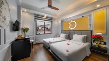 Habitación de hotel con dos camas, ventilador de techo, televisor, ventana con vista a la ciudad y un florero decorativo con flores.