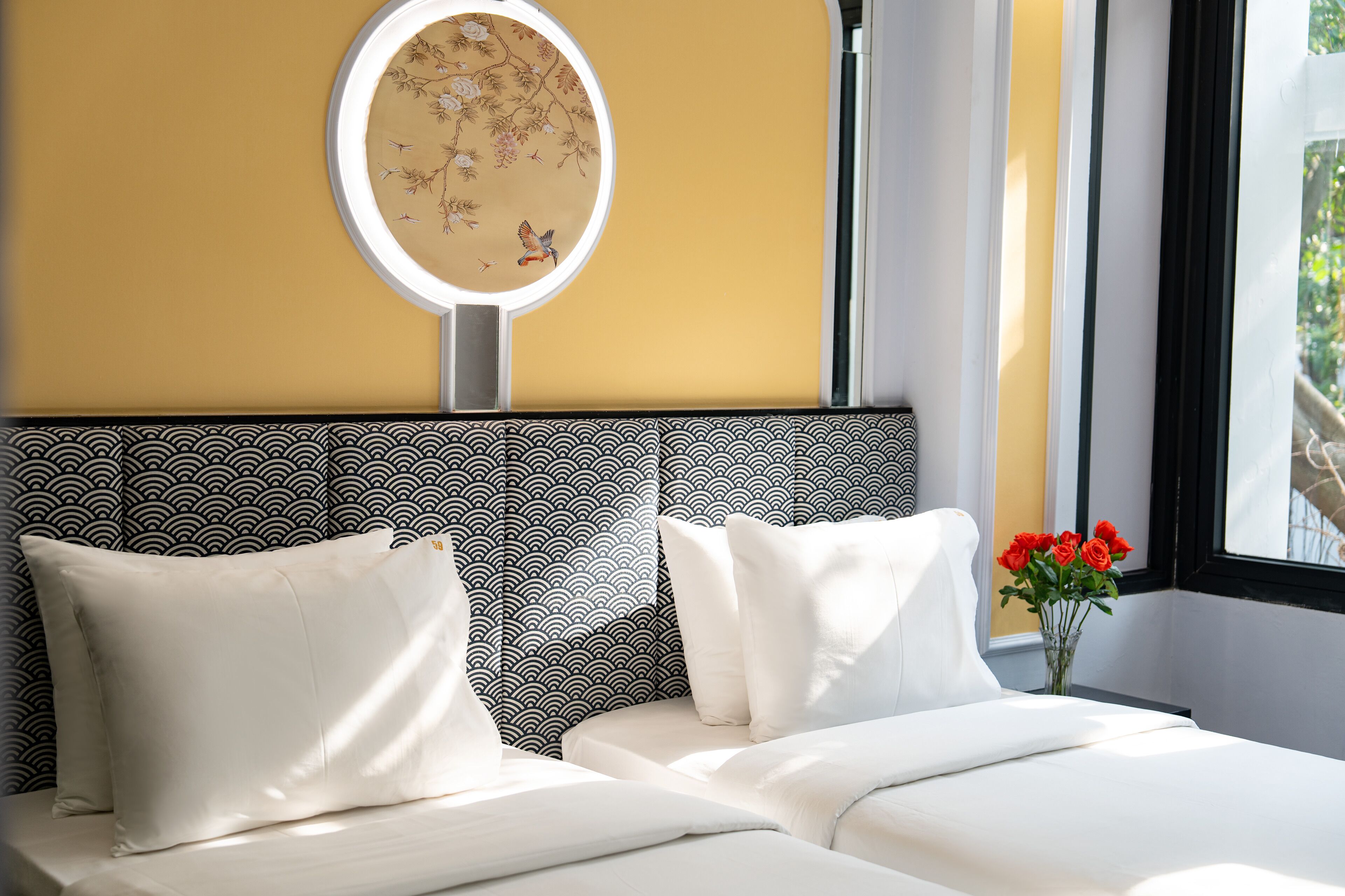A hotel room with a bed, pillows, a round mirror, and a vase with flowers.