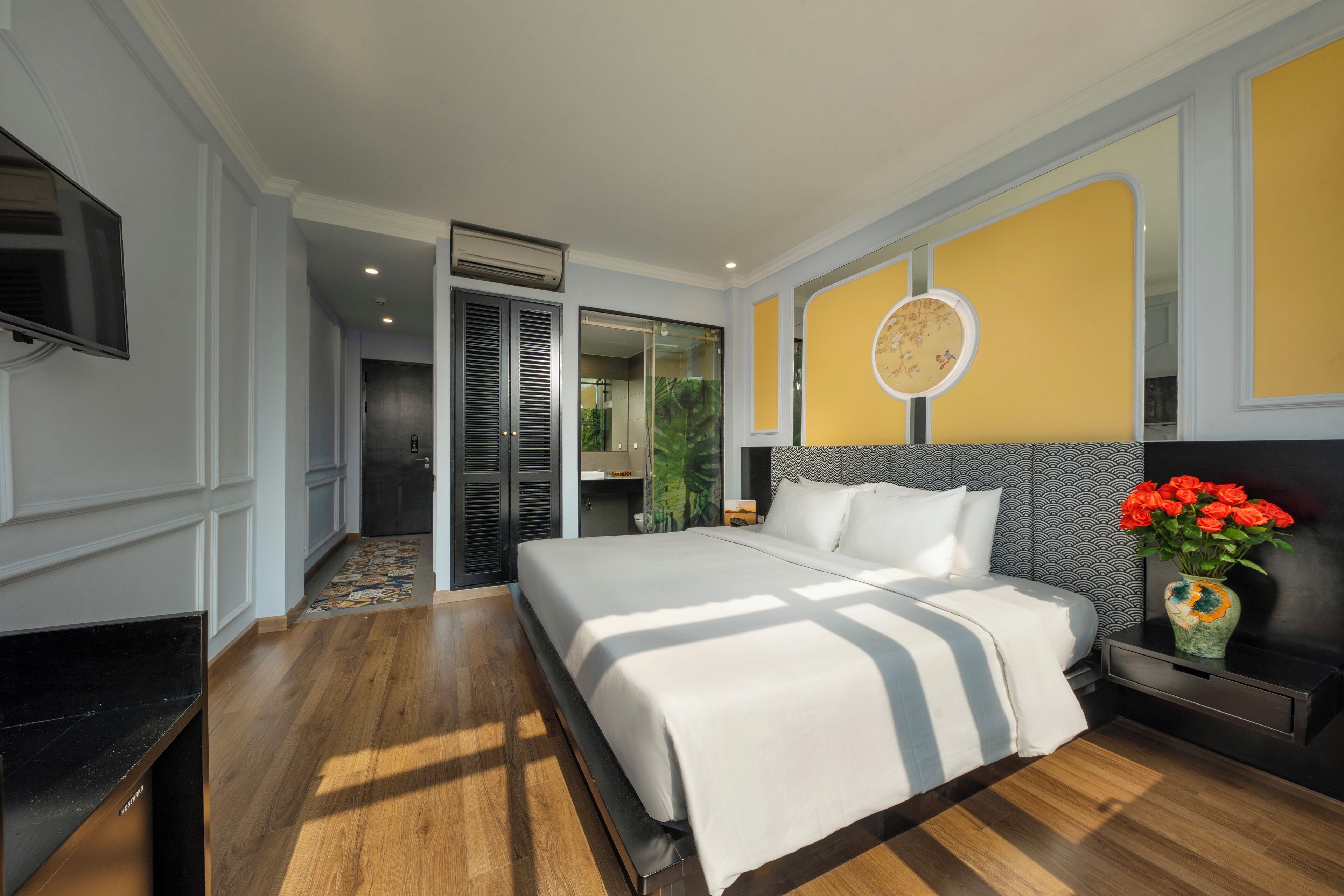 A modern hotel room with a large bed, a TV, a small table with a vase of flowers, and a sliding door leading to a bathroom.