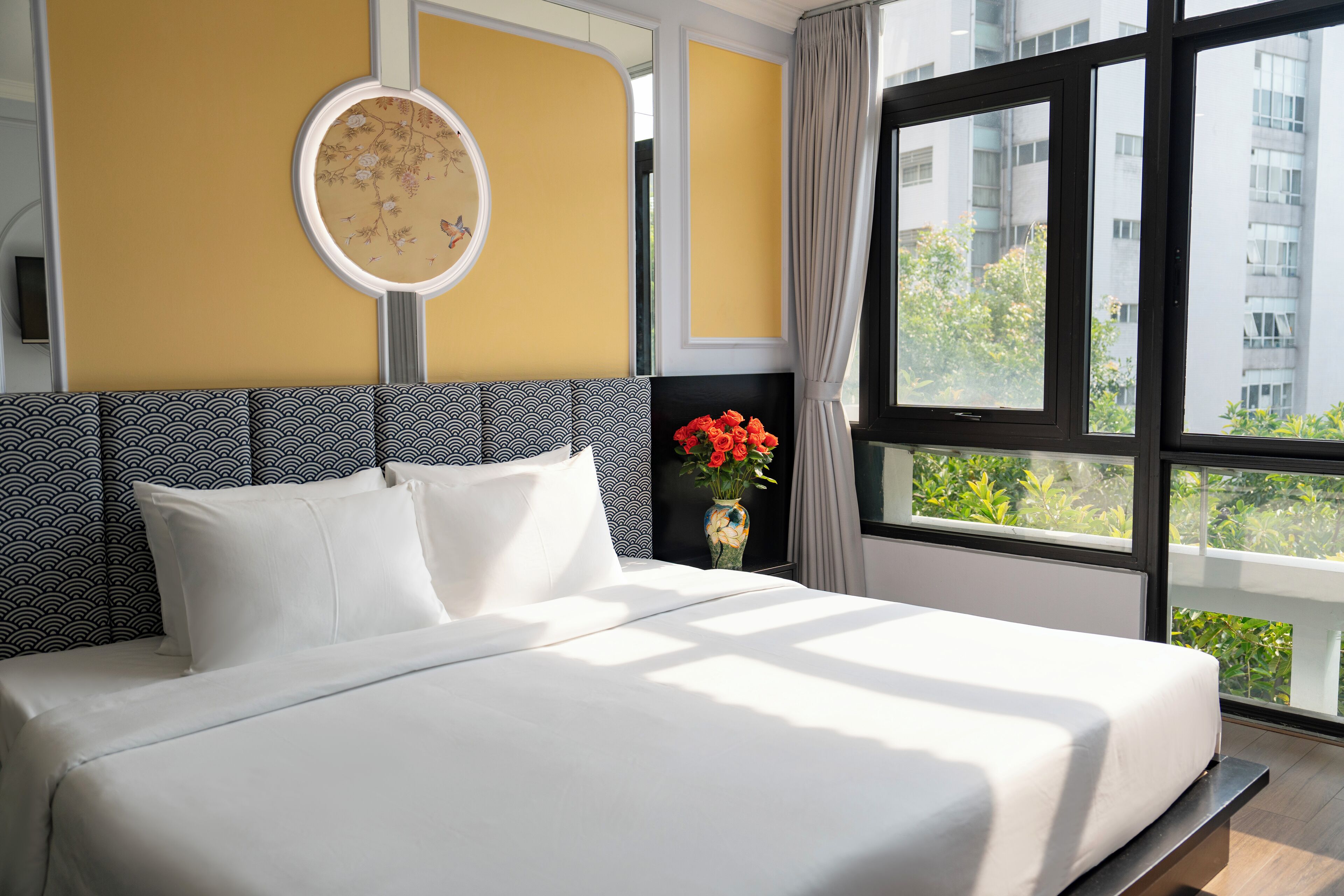 A hotel room with a large bed, a round mirror on the wall, and a view of greenery outside.