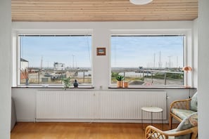A room with wooden flooring, two windows with a view of boats, a radiator, a small table, and a wicker chair.
