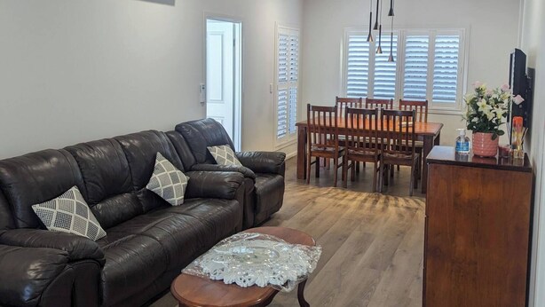 A spacious living room with a leather sofa, a wooden coffee table, and a dining area with a wooden table and chairs. There are ceiling fans and an air conditioning unit. - Immaculate Entire House in Chermside West 2-Beds (Chermside West)