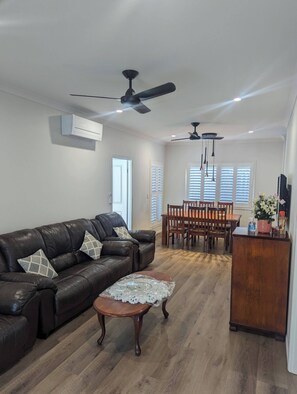 A spacious living room with a leather sofa, a wooden coffee table, and a dining area with a wooden table and chairs. There are ceiling fans and an air conditioning unit.
