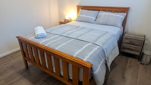 A single bed with a wooden frame and a white bedspread, a bedside table with a lamp, and a trash bin. - Immaculate Entire House in Chermside West 2-Beds (Chermside West)