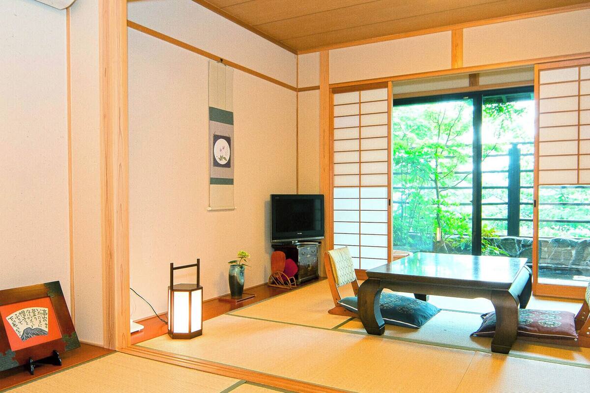 Ein traditioneller japanischer Raum mit Tatami-Böden, einem niedrigen Tisch, einem Fernseher und einer Schiebetür mit Blick auf Grünflächen.