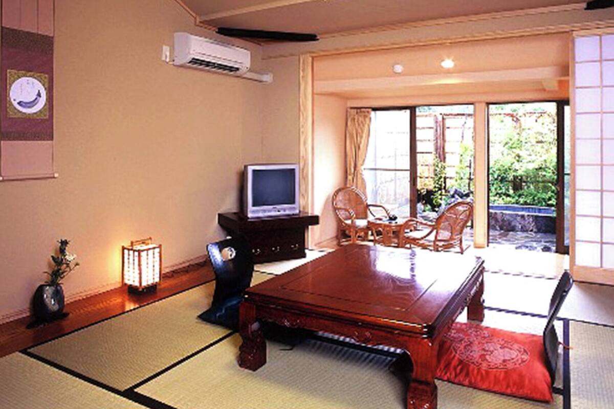 Ein traditioneller japanischer Raum mit Tatami-Böden, einem Fernseher auf einem Schrank und einer Schiebetür zu einem Außenbereich.