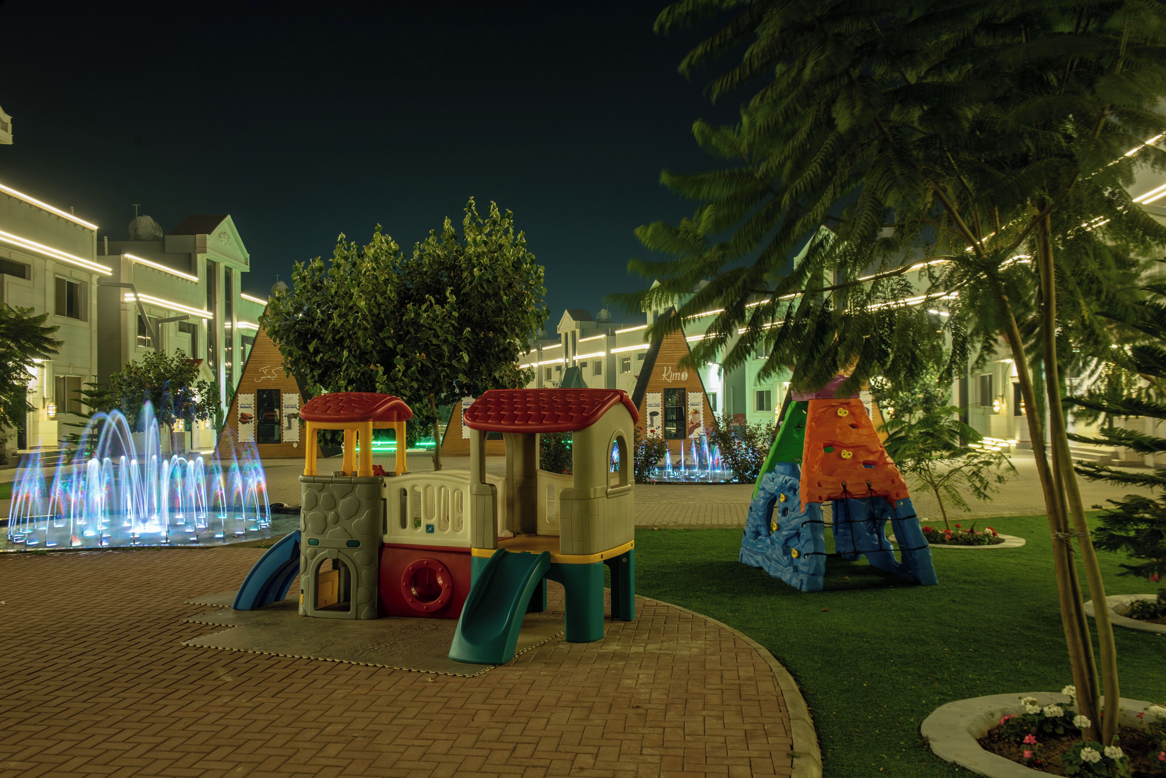 Zona de juegos infantiles con un tobogán y estructura para escalar, rodeada por un sendero de ladrillos e iluminada con luces nocturnas.
