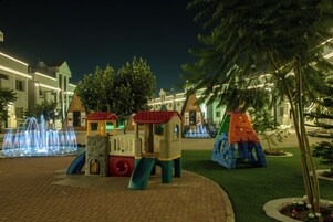 Zona de juegos infantiles con un tobogán y estructura para escalar, rodeada por un sendero de ladrillos e iluminada con luces nocturnas.