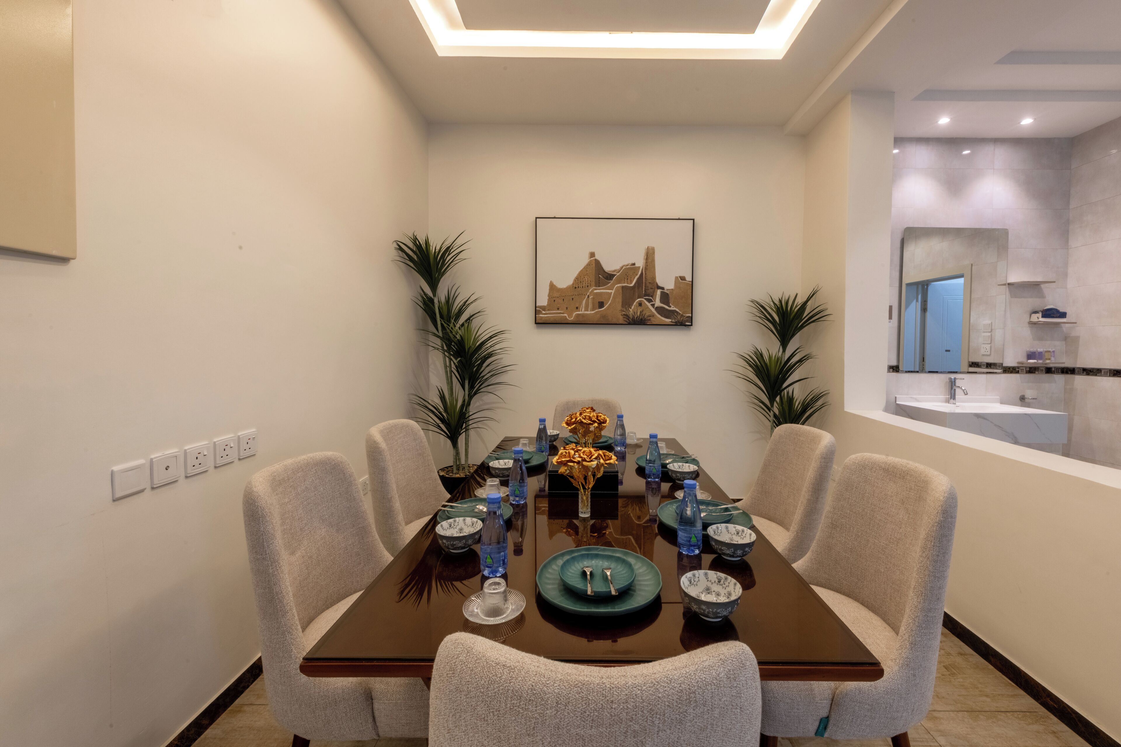 A dining area with a rectangular table set for four, surrounded by chairs. There is a framed picture on the wall above the table.