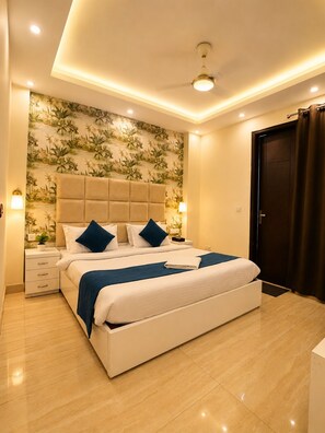 A modern bedroom with a large bed, a headboard, bedside lamps, a nightstand, and a door with curtains. - Ambient Express Cr Park (New Delhi)